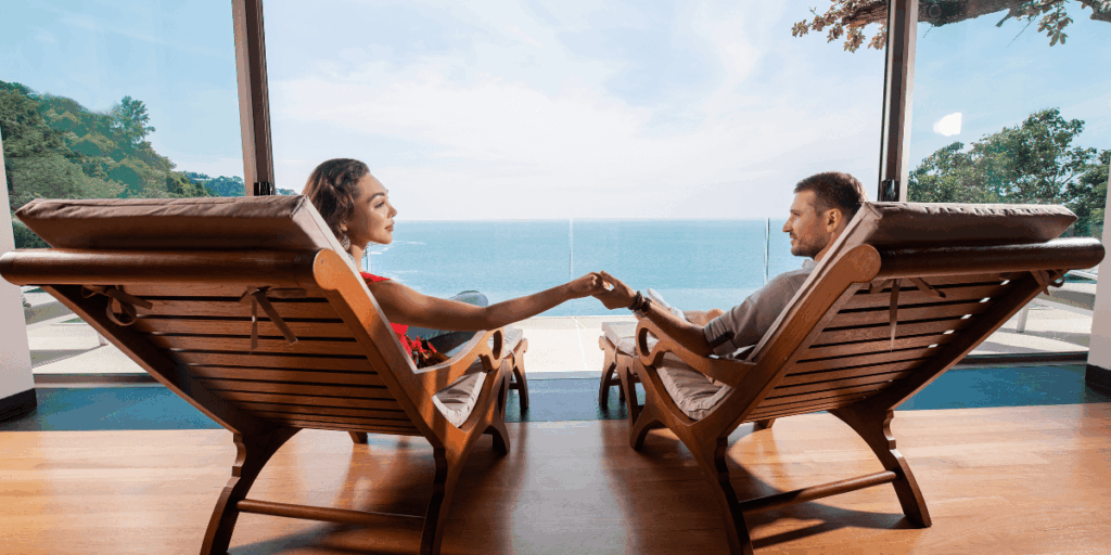 A man and woman sit in chairs, enjoying the ocean view in the background on a sunny day.