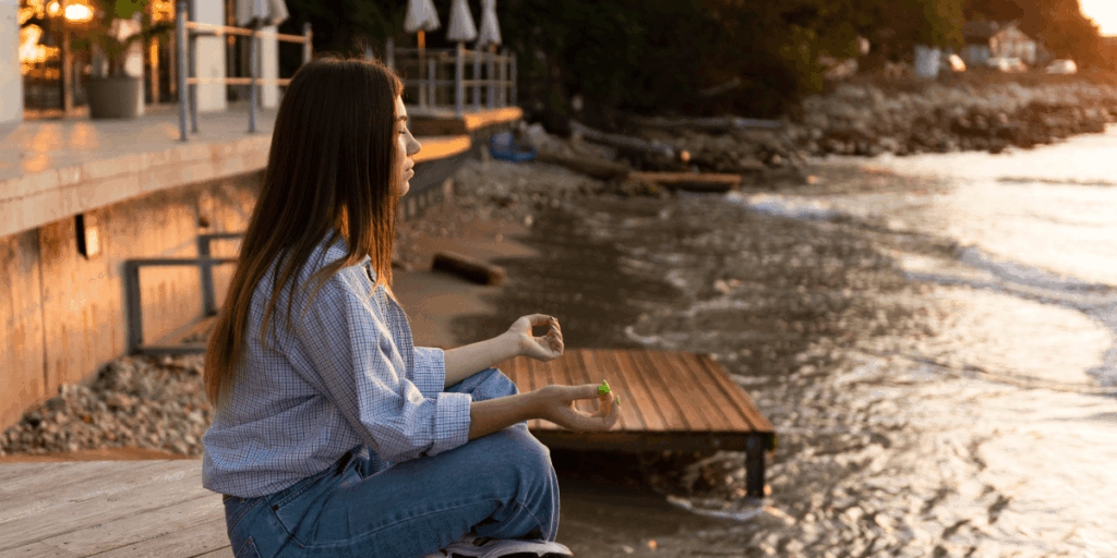 A woman sits cross-legged on the beach, meditating as the sun sets, casting warm hues across the sky and ocean.